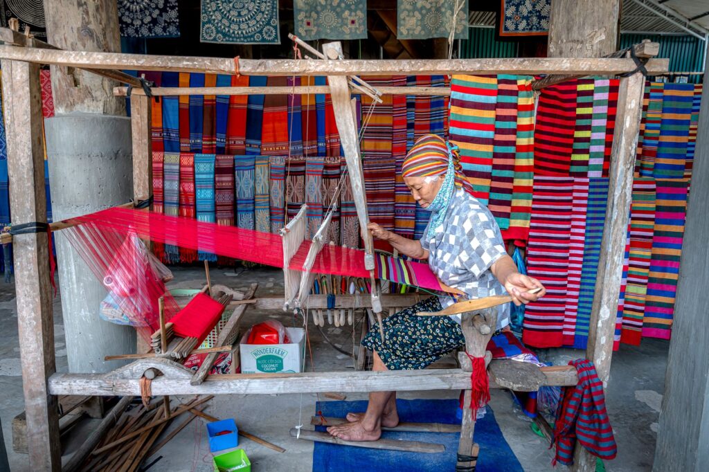 Elderly woman weaving colorful fabrics on a loom at a vibrant village market, emphasizing traditional craft.