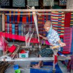 Elderly woman weaving colorful fabrics on a loom at a vibrant village market, emphasizing traditional craft.