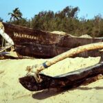 A wooden fishing boat named 'Vailankanni' rests on a sandy beach in India, surrounded by lush trees.