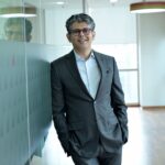 Smiling businessman standing confidently in a modern office hallway, wearing a suit.