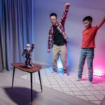 Father and son enjoy dancing with a humanoid robot in a colorful home setting.