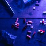 A flat lay of various drugs including pills, capsules, and a syringe on a wooden surface under moody blue lighting.