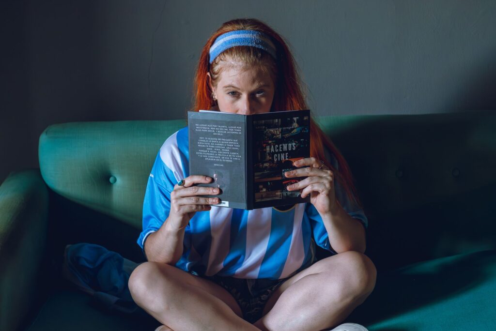 A young woman reads a book on a sofa, wearing an Argentina soccer jersey indoors.