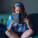 A young woman reads a book on a sofa, wearing an Argentina soccer jersey indoors.