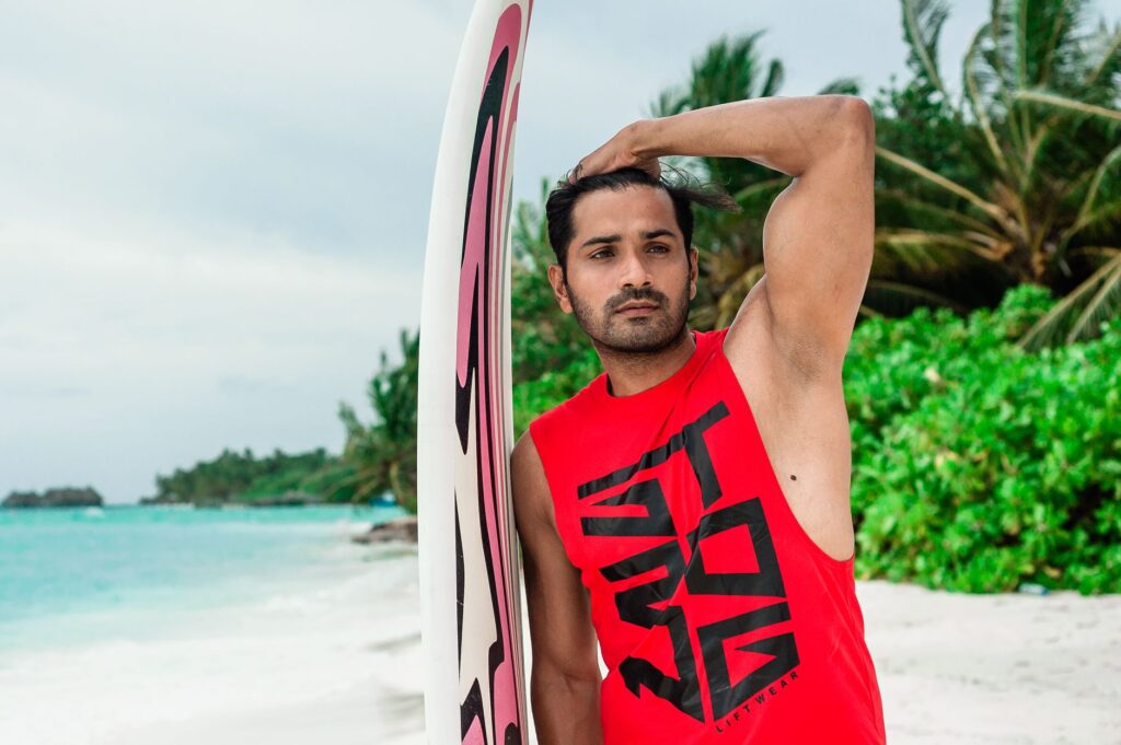 A male model poses with a surfboard on a tropical Maldives beach, showcasing summer fashion.