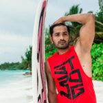 A male model poses with a surfboard on a tropical Maldives beach, showcasing summer fashion.