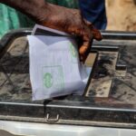 A close-up shot of a hand placing a paper ballot into a transparent voting box outdoors.