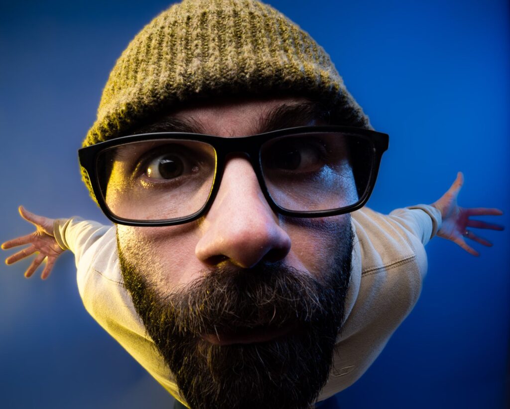 Close-up fisheye portrait of a bearded man wearing glasses and a beanie against a blue background.