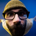 Close-up fisheye portrait of a bearded man wearing glasses and a beanie against a blue background.