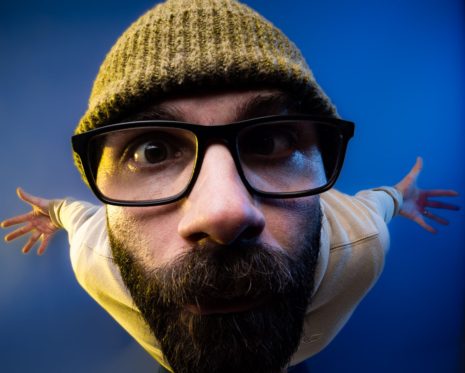 Close-up fisheye portrait of a bearded man wearing glasses and a beanie against a blue background.