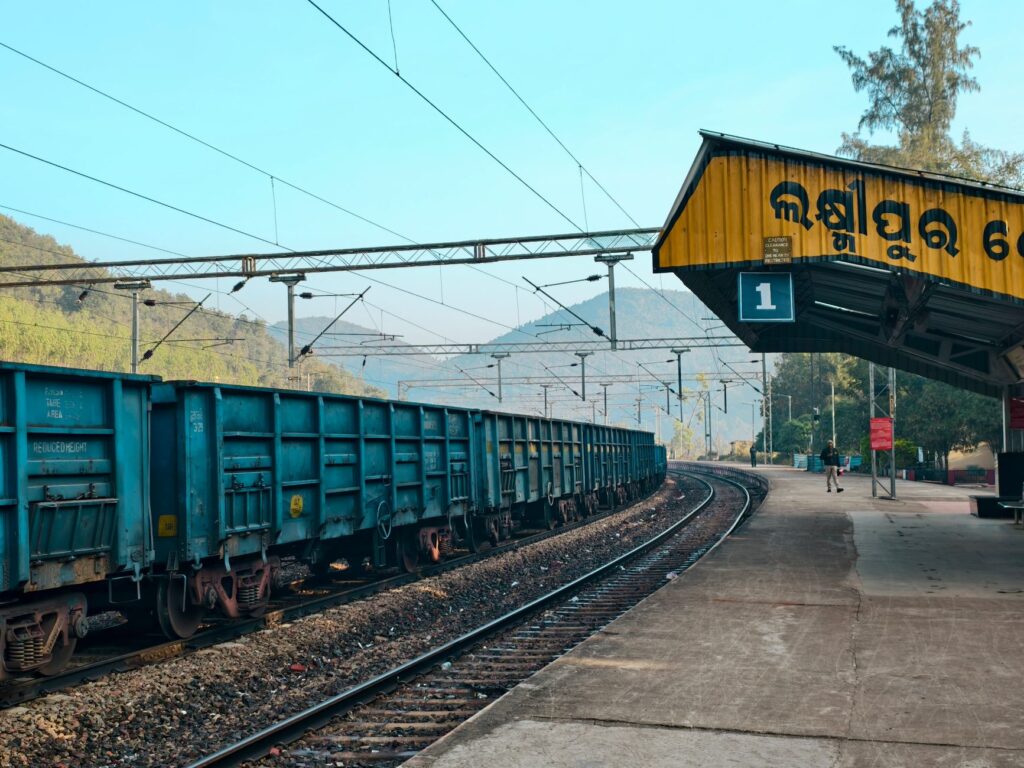 A blue freight train at an Indian railway station platform with mountains in the background.