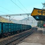 A blue freight train at an Indian railway station platform with mountains in the background.