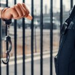 Close-up of a police officer handcuffing an individual against a metal fence outdoors.