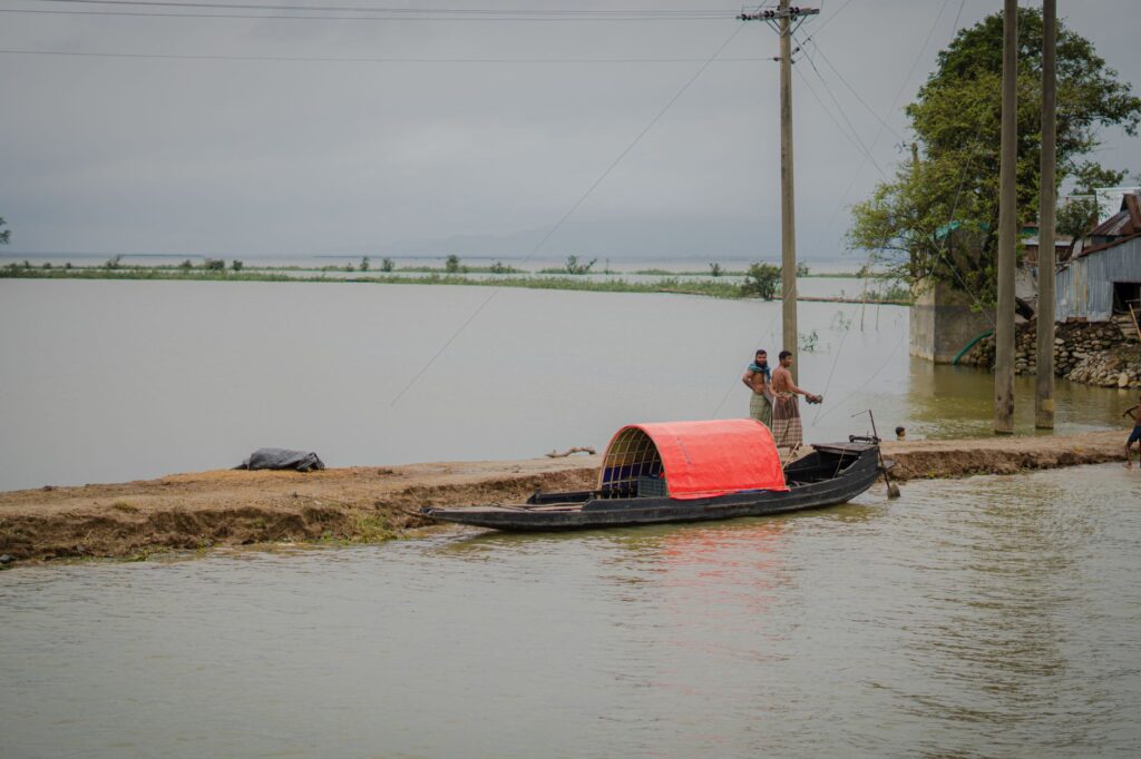 Two men stand beside a traditional canoe on a flooded riverbank under cloudy skies.