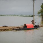 Two men stand beside a traditional canoe on a flooded riverbank under cloudy skies.