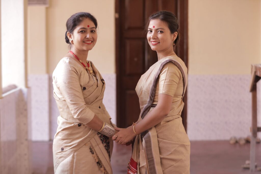 Two South Asian women in traditional sarees smiling indoors, reflecting Indian cultural attire.