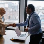 Young female candidate shaking hands with employer before job interview in light office with urban landscape through windows