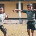Two boys happily play outside a rural school in Assam, India, showcasing the joy of childhood and friendship.