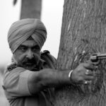 Black and white image of a police officer in uniform aiming a revolver behind a tree.