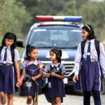 Schoolgirls in uniforms walk safely alongside a police escort on a rural road.