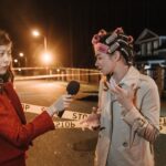 Asian female journalist interviews a woman at a night crime scene. Police tape marks the area.