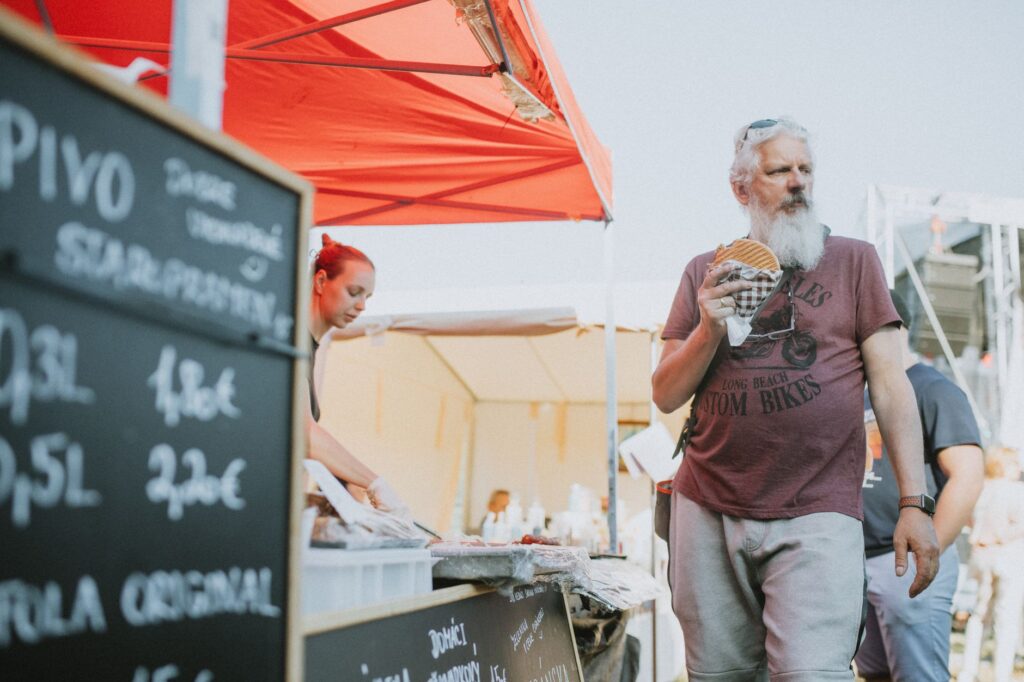 Man eating a burger at a street food market with beer menu in view, vibrant street scene.