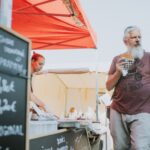 Man eating a burger at a street food market with beer menu in view, vibrant street scene.
