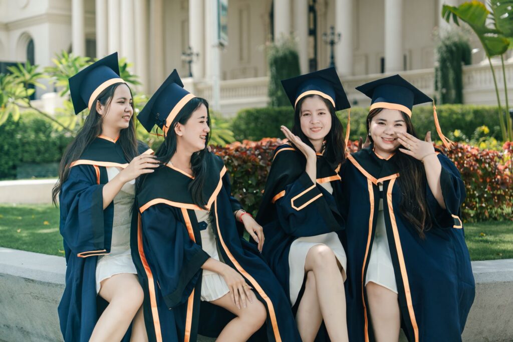 Group of four happy female graduates celebrating together in caps and gowns outdoors.