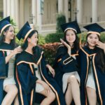 Group of four happy female graduates celebrating together in caps and gowns outdoors.
