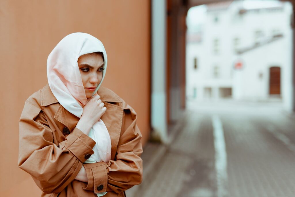 A Muslim woman in stylish clothing stands in an urban setting with a thoughtful expression.
