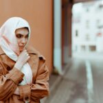 A Muslim woman in stylish clothing stands in an urban setting with a thoughtful expression.