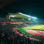 A crowded night-time football match in a brightly lit and bustling stadium, full of energy.