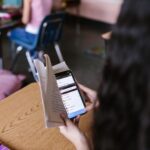 A student in a classroom multitasks by reading a book while using a smartphone, emphasizing modern educational challenges.
