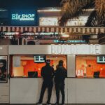 Two people standing at a box office under neon lights at night, urban cinema setting.