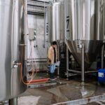 An industrial worker inspects fermentation tanks in a brewery, surrounded by equipment and machinery.