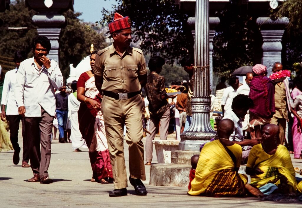 A bustling street scene in India featuring a diverse crowd and a policeman walking amidst people.