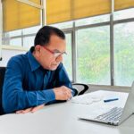 Focused man in blue shirt works intently on a laptop in a bright office setting.
