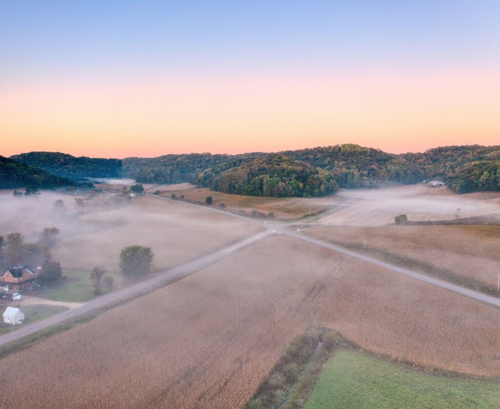 A scenic aerial view of misty farmland and rolling hills during sunrise in Urne, Wisconsin.