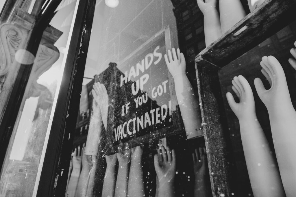 Monochrome image of mannequin hands in a store window displaying 'Hands Up if You Got Vaccinated!' signage.