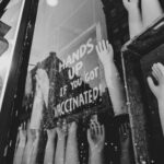 Monochrome image of mannequin hands in a store window displaying 'Hands Up if You Got Vaccinated!' signage.