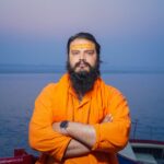 A spiritual Hindu sadhu stands confidently by the Ganges River in Varanasi, India, during twilight.