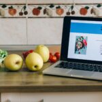 Laptop showing a delivery app beside fresh fruits and vegetables on a kitchen counter.