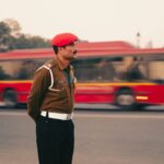 A traffic officer in a red beret stands in New Delhi, with a blurred red bus in the background.