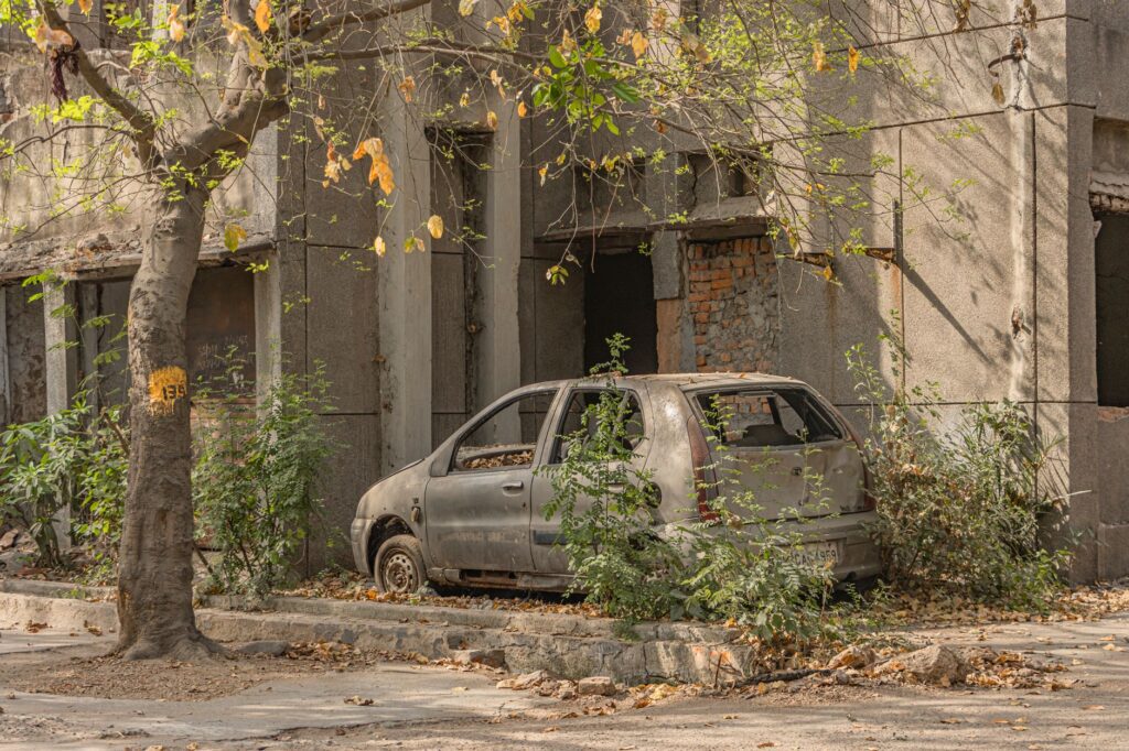 An old, abandoned car parked next to a dilapidated building amidst foliage in Delhi, India.