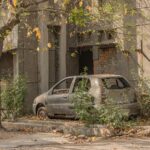 An old, abandoned car parked next to a dilapidated building amidst foliage in Delhi, India.