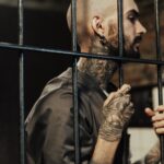 A tattooed man behind prison bars, contemplating life inside a penitentiary.