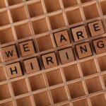 Wooden letter tiles arranged to spell 'We Are Hiring' on a wooden grid background.