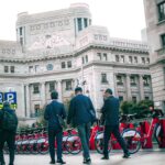 Businessmen walk past BBVA building in city with rental bikes.