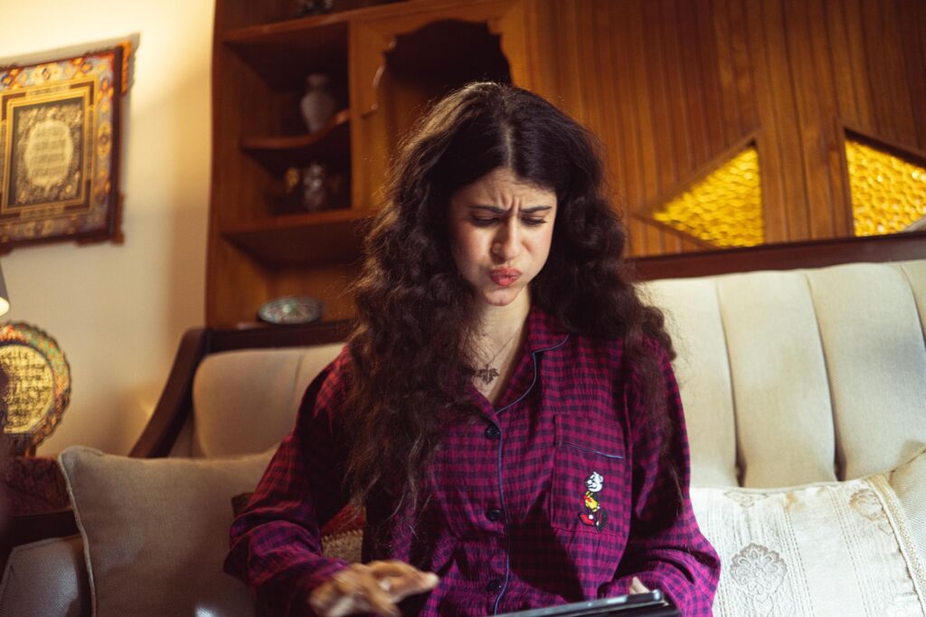 A woman sitting indoors looking frustrated with a tablet in hand, wearing pajamas.