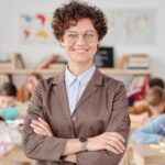 Smiling teacher with glasses standing with arms crossed in a classroom, students learning.